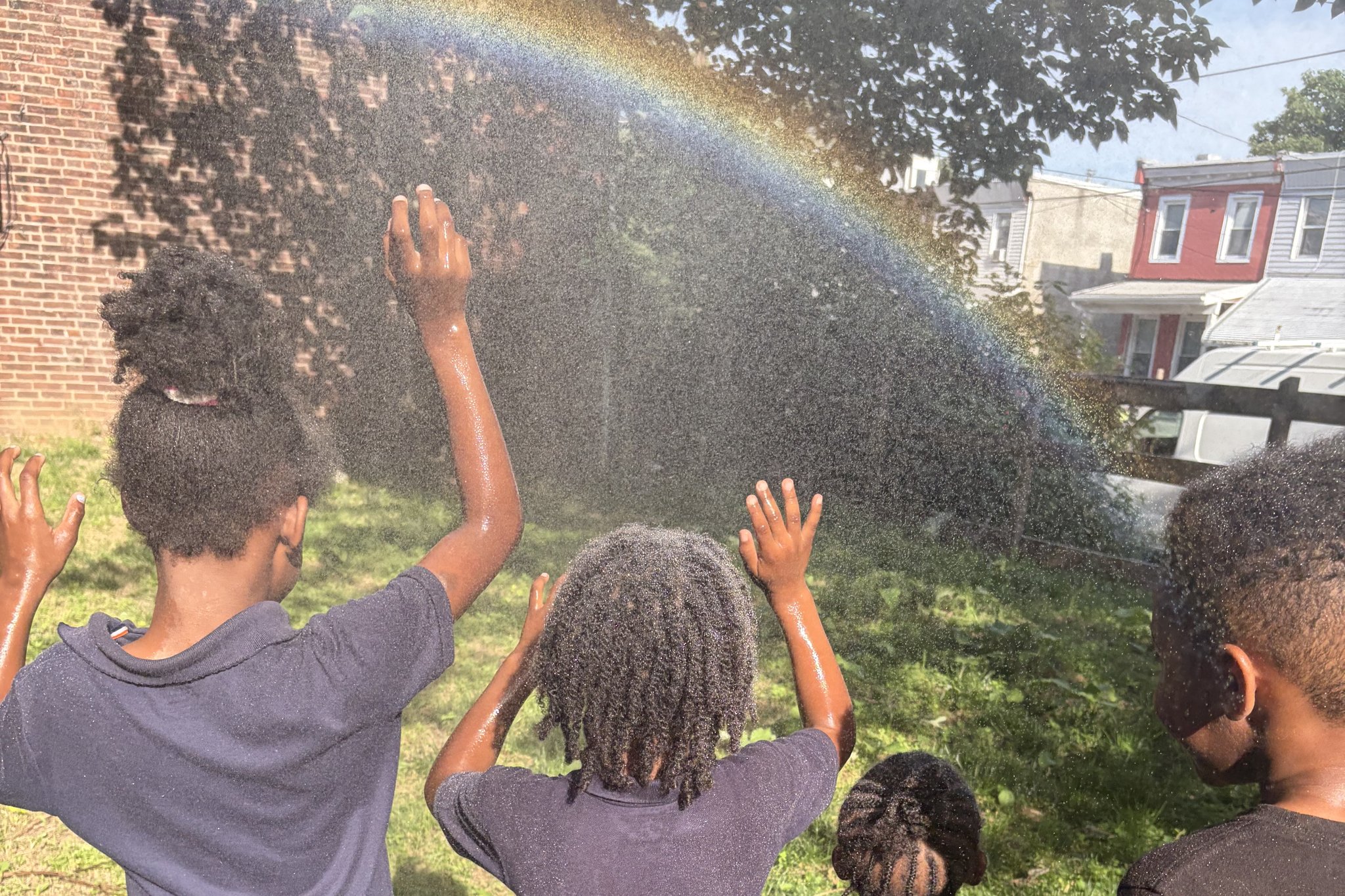 Children raise their hands under a rainbow of water spray in a neighborhood green space.